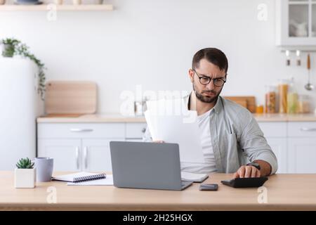 Busy adult caucasian man with beard in glasses counts finances, pays bill and taxes for calculator Stock Photo
