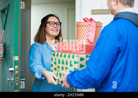 Delivery man bringing holiday packages. Woman at home standing in doorway, receiving parcels for Christmas gifts. Stock Photo