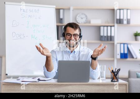 Happy adult european man teacher in glasses, headphones spreads arms to sides and looks at laptop Stock Photo