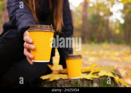Girl sitting on a tree stump holding out one of two coffee cups closer to the camera. Coffee break in the autumn forest. Stock Photo