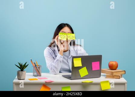 A lot of work concept. Tired asian female student sleeping with stickers on eyes, sitting at table over blue background Stock Photo