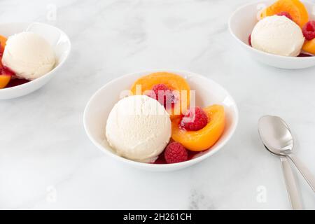 Vanilla ice cream with peaches, raspberries and raspberry sauce in bowls on white marble with copy space. Low angle view. Stock Photo