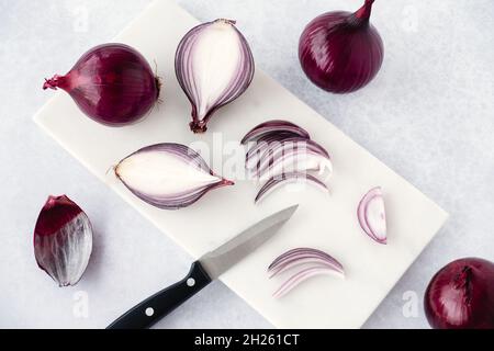 Red raw onions, whole, halved, quartered and sliced on white marble chopping board with knife. High angle view and copy space. Stock Photo