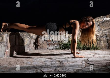 blonde woman dressed in black doing push-ups at night with her feet on a wall Stock Photo