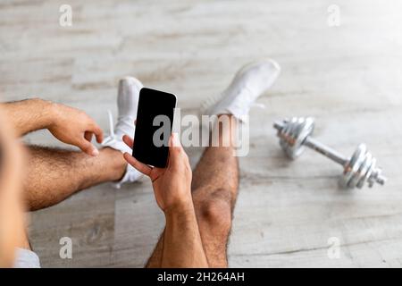 Cropped view of fit young guy with dumbbell checking new app on mobile phone during break, mockup for design Stock Photo