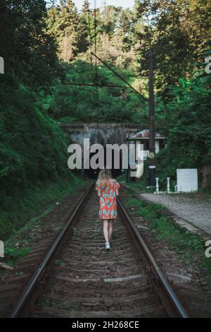 Young woman in colourful dress walks along the rails in the forest. Lonely girl approaches a tunnel in the green forest. Back side view. Stock Photo