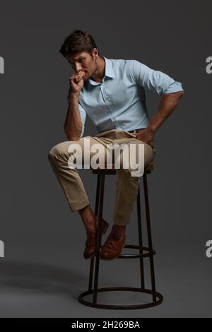 Young thoughtful and worried european businessman sitting on bar chair. Bearded man with dark hair wear shirt with trousers. Isolated on dark gray bac Stock Photo