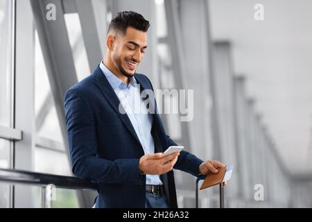 Mobile Roaming. Young Smiling Middle Eastern Businessman Using Smartphone In Airport Stock Photo