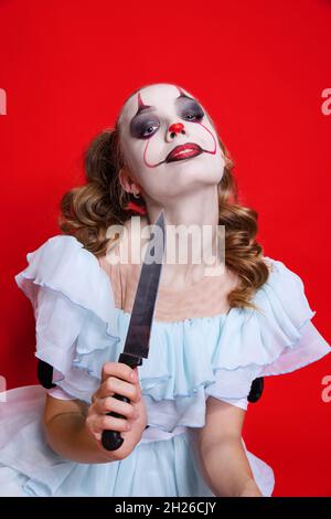 Halloween spooky theme. Mystical blonde girl with bright holiday make up holding knife isolated over red background Stock Photo
