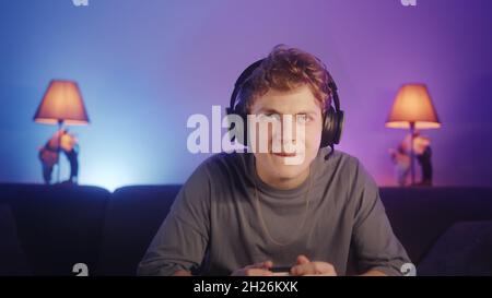 Handsome blonde-hair young gamer boy sitting on couch and playing game on a console excitedly. He wearing headphones, plays with a wireless controller Stock Photo