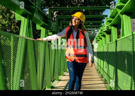 Beautiful young female engineer or supervisor, wearing safety equipment, inspects a railroad pedestrian bridge. concept of empowered woman, profession Stock Photo