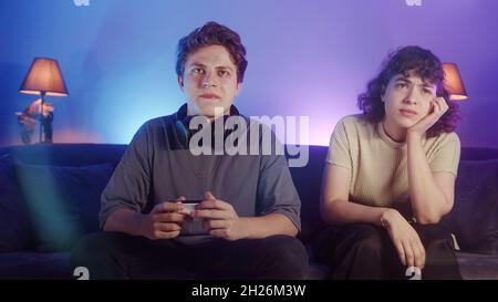 Young curly hair girl offended to her careless lover playing video games with wireless controller at home. Indifferent young blonde man having gaming Stock Photo