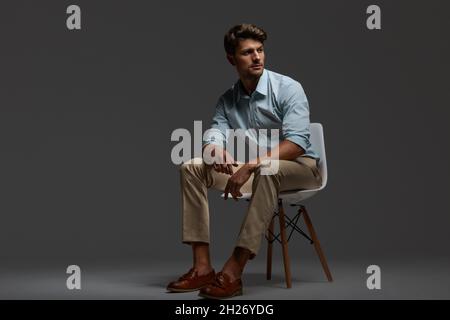 Young beautiful concentrated european businessman sitting on chair. Bearded man with dark hair wear shirt with trousers. Isolated on dark gray backgro Stock Photo