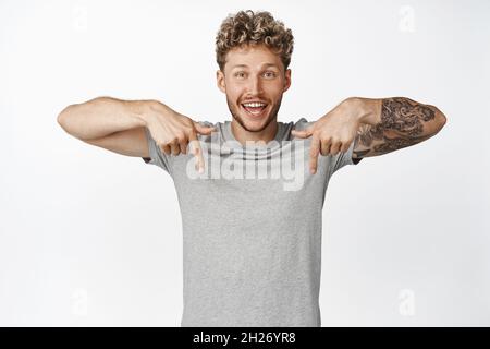 Portrait of handsome smiling blond man pointing fingers down, showing advertisement, making announcement below, standing over white background Stock Photo