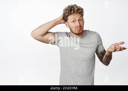 Clueless young man scratch his head and shrugs confused, dont know understand, has no idea, stands puzzled against white background Stock Photo