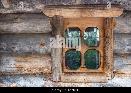 Small window on the wall of a log house, the reflection of green trees in the glass. Stock Photo