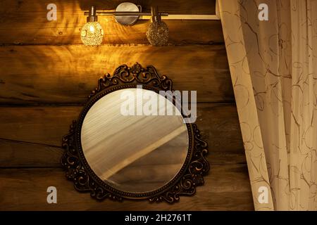 Decorative-framed round mirror hangs on a wooden wall under a lamp next to a curtain. Stock Photo