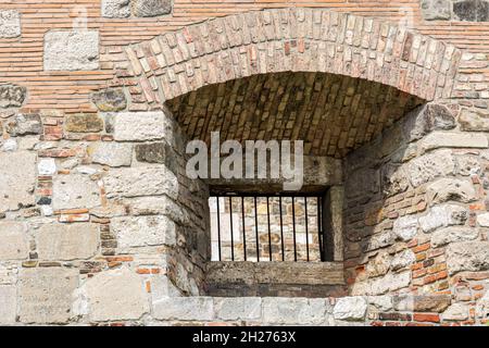 Window with a lattice of iron rods in a depression of a thick stone fortress wall with a brick arch. Stock Photo