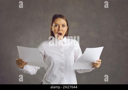 Woman holding paper documents and looking at camera with shocked face expression Stock Photo