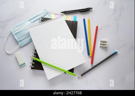 Blank spiral notepad mock up with copy space, crayons, surgical face mask against covid-19 infection and stationary supplies on a marble surface, high Stock Photo