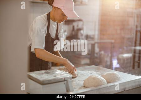 Woman in pink cap kneads raw dough piece for bread in craft bakery shop Stock Photo