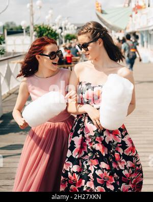 Close up portrait of smiling excited girls with sunglasses and prom dresses holding cotton candy at amusement park. Teenagers in pink evening dresses Stock Photo