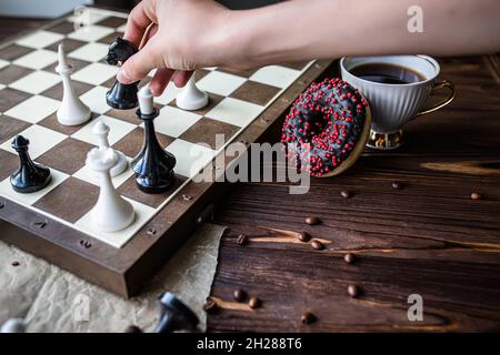 hand holds a white vintage cup of coffee. Chess board and scattered chess on wooden table. Chocolate doughnut for morning breakfast. board game Concep Stock Photo