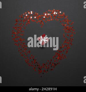A miniature red box in the middle of a shiny heart lined with canfetti stars on a gray background Stock Photo