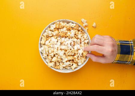 Male hand picking popcorn from a bowl, yellow background, top view. Movie entertainment concept Stock Photo