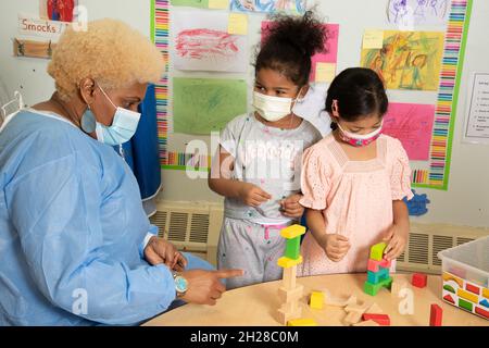 Education Preschool 4-5 year olds two girls building with colorful wooden blocks, teacher in protective gown talking to them about their work Stock Photo