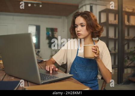Serious female owner of small business sitting at table and using laptop while drinking coffee and working in pottery workshop Stock Photo