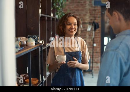 Smiling attractive clerk in apron standing at shelf and selling ceramic cup to man in pottery store Stock Photo