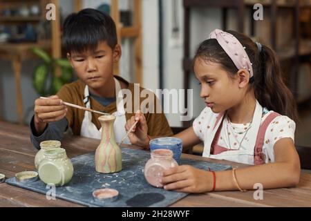 Concentrated interracial kids sitting at wooden table and painting clay vase together in workshop Stock Photo