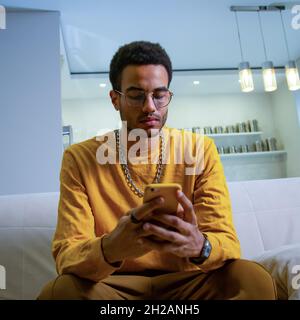 A young, happy African man smiles and texts on his smartphone. Happy black man using smart phone at home Stock Photo