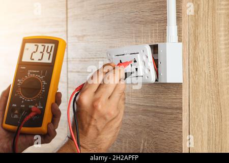 Electrician using a digital meter to measure the voltage at a wall socket on a wooden wall. Stock Photo