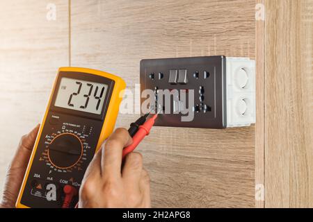 Electrician using a digital meter to measure the voltage at a wall socket on a wooden wall. Stock Photo