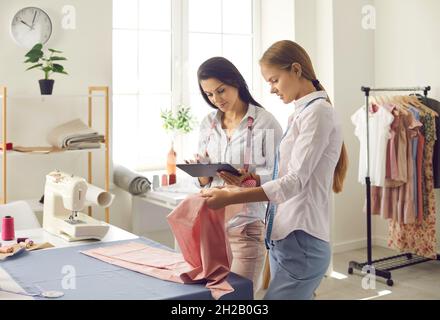 Two female atelier owners check catalog with new fabric samples on digital tablet. Stock Photo