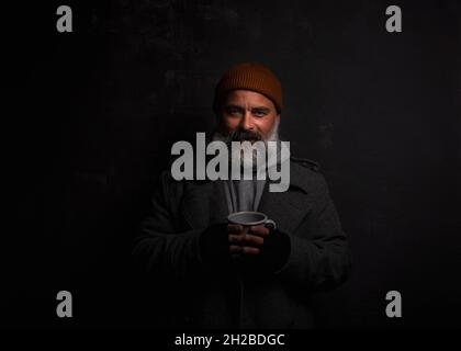 Bearded beggar man smiling, happy even living on the street. Cheerful homeless man with cup of hot tea isolated on dark background Stock Photo