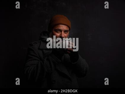 Bearded beggar man smiling joyfully with cup of hot tea isolated on black background Stock Photo