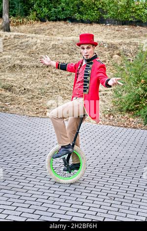 Full body man in costume and hat stretching out arms and balancing while riding unicycle on pavement during performance in park Stock Photo