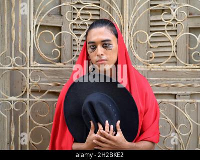Handsome young nonbinary Latino covers his head with a red Mexican rebozo and holds a black hat in front of a white wrought iron window grill. Stock Photo