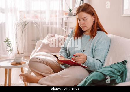 Home woman taking notes in notebook while using tablet gadget at home. Freelance writing details on book while working on in living room. Stock Photo