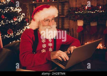 Photo of cheerful glad santa claus look sit workplace work laptop wear hat pullover in north pole office indoors Stock Photo