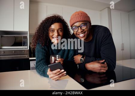 Multi-ethnic couple smiling at camera while in kitchen in modern apartment using cellphone Stock Photo