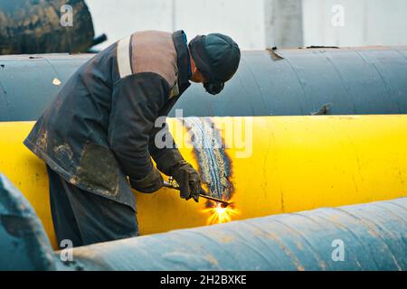 The welder cuts a large yellow pipe with acetylene welding for gasification. Disposal of old used metal pipes. Authentic workflow scene. Industrial background. Stock Photo