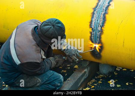 The welder cuts a large yellow pipe with acetylene welding for gasification. Disposal of old used metal pipes. Authentic workflow scene. Industrial background. Stock Photo