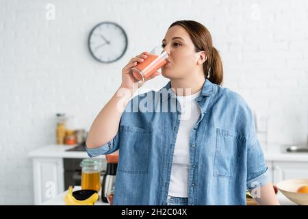 Plus size woman drinking fruit smoothie in kitchen Stock Photo