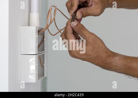 The electrician is using a knife to cut the wire to install the power switch in the box on the wall. Stock Photo