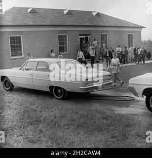 1960s, historical, outside a single-storey building, housing a business college, with students and teachers looking on, a teenage girl in a skirt and ankle socks using a jet spray to wash an American Chevrolet automobile, Middletown, Virginia, USA. Stock Photo