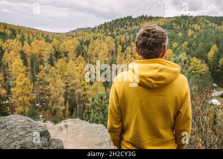 A man in a yellow hoodie stands on top of a mountain, admiring the magnificent landscape of the autumn forest from above. Sustainable environment.
